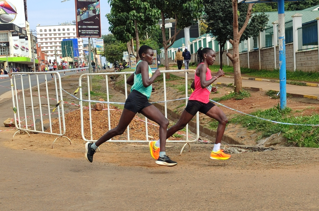 Two runners in a street race sprint past metal barriers along a city sidewalk, wearing turquoise and pink tops.